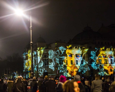 Na zdjęciu w centralnej części na całej powierzchni fragment głównego budynku Teatru. Fasada jest oświetlona. Kolorowe światła przedstawiają twarz Wyspiańskiego. Twarz jest rozłożona na całym fragmencie fasady. Pojawia się cyklicznie jedna obok drugiej. Na pierwszym planie grupa ludzi patrząca w kierunku budynku. Stoją tyłem. Tło jest czarne.