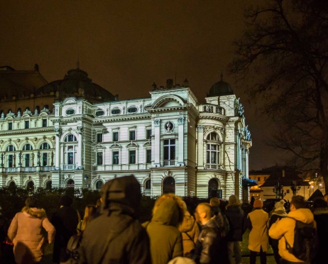 Zdjęcie zrobione z plant. Duża liczba osób stoi na chodniku i patrzy w stronę budynku Teatru. Budynek oświetlony jest na w trójwymiarze w kolorze białym. Po prawej stronie w oddali jest drzewo bez liści lekko zachodzące na prawą stronę budynku. Tło jest czarne.