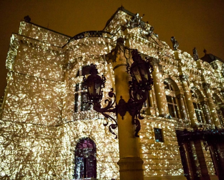 Na zdjęciu fragment głównego budynku Teatru. Na pierwszym planie zgaszona zdobiona latarnia. Za nią oświetlona ściana frontowa budynku ujęcie z boku żółto złotym światłem. Przypomina to zapalone malutkie światełka. Tło jest ciemne.