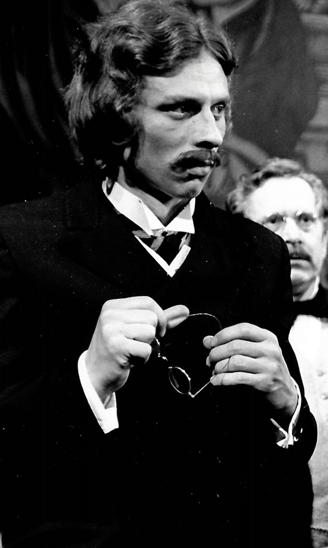 Czarno-białe zdjęcie aktora Mikołaja Grabowskiego z lat
młodości. Widoczny do pasa, ubrany w surdut i białą koszulę. W rękach trzyma
okulary. Włosy półdługie, lekko falowane, wąsy. Patrzy w bok.