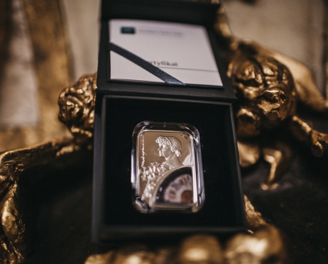 Na zdjęciu moneta z podobizną Heleny Modrzejewskiej. Srebrna, prostokątna w prawym dolnym rogu figury kolorowy wachlarz. Moneta umieszczona jest w granatowym futerale. Pudełko leży na fragmencie złotej rzeźby.