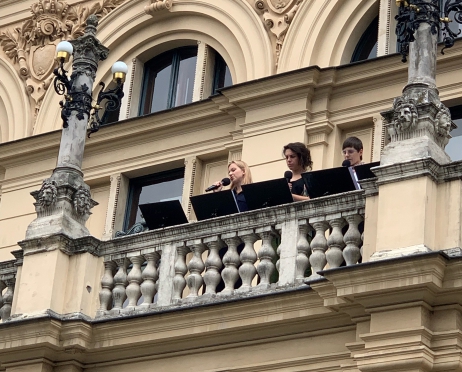 Na zdjęciu fragment budynku Teatru im. Juliusza Słowackiego w Krakowie. Budynek jest jasny, zdobiony rzeźbionymi głowami. Na jednym z balkonów znajdują się aktorzy. Trzy młode kobiety stoją przed pulpitami i czytają do mikrofonów.