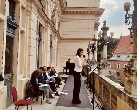 Na zdjęciu grupa osób siedzi na balkonie Teatru im. Juliusza Słowackiego w Krakowie. Jedna z kobiet stoi na podeście i czyta do mikrofonu. Pozostali siedzą na krzesłach ustawionych wzdłuż ściany budynku. Na końcu balkonu znajduje się fortepian przy którym siedzi muzyk.