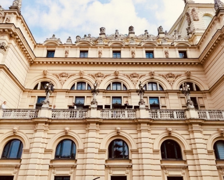 Na zdjęciu fragment budynku Teatru im. Juliusza Słowackiego w Krakowie. Budynek jest jasny, zdobiony rzeźbionymi głowami. Na jednym z balkonów znajdują się aktorzy.