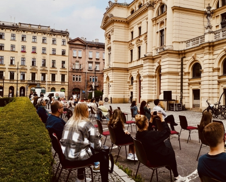 Na zdjęciu grupa osób siedzi na krzesłach na parkingu Teatru im. Juliusza Słowackiego w Krakowie. Krzesła ustawione są w czterech rzędach, nie wszystkie są zajęte. Widzowie zwróceni są w stronę jednego z balkonów budynku, na którym stoją aktorzy.