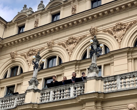 Na zdjęciu fragment budynku Teatru im. Juliusza Słowackiego w Krakowie. Budynek jest jasny, zdobiony rzeźbionymi głowami. Na jednym z balkonów znajdują się aktorzy. Dwie kobiety i dwaj mężczyźni stoją przed pulpitami i czytają do mikrofonów.