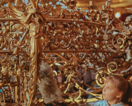 Zbliżenie na fragment ogromnego, bogato zdobionego, złotego żyrandola. Przy nim stoi dziewczynka, około 10 lat, włosy w kucyk, ubrana w siwą bluzę z kapturem w kwiatki. W ręku trzyma miotełkę do kurzu, dotyka nią żyrandola. Jest skupiona.