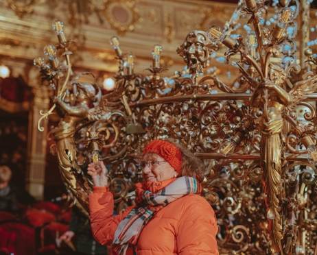 Fragment wielkiego złotego żyrandola opuszczonego na widownię Teatru Słowackiego. Obok stoi kobieta pozująca do zdjęcia obok. Stoi nieruchomo, trzyma rękę na żarówce wkręconej w żyrandol. Ubrana jest w puchową pomarańczową kurtkę, owinięta szalem w kratę, na głowie ma pomarańczową wełniana opaskę. Włosy brązowe, kręcone do ramion i grzywkę. przez ramię przewieszona wielka zielona torebka. kobieta uśmiecha się. W tle loże teatralne.