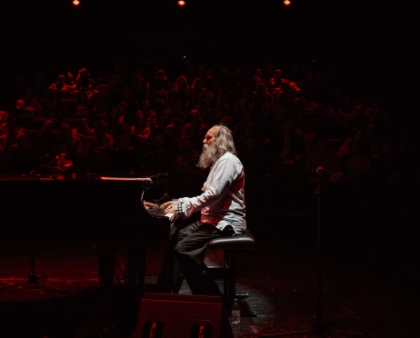 Na zdjęciu mężczyzna siedzi bokiem. Gra na fortepianie. Patrzy prosto przed siebie. Światło jest skierowane tylko na niego. Ubrany jest w białą bluzkę i szare spodnie.Tło jest czarno-czerwone.