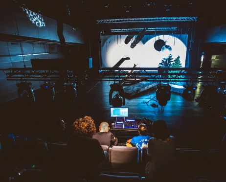 Ujęcie na scenę teatralną z widowni. Tyłem na krzesłach widoczne kilka postaci. Przed nimi przed pierwszym rzędem krzeseł stoi oświetlona konsola. Nad scenę opuszczony jest sztankiet z przymocowanymi do niego reflektorami. Na scenie leży kilka elementów: bal drzewa, krzew, betonowe płyty, zjeżdżalnia. Na tylnej ścianie sceny wielki ekran, na którym widoczne są dwa białe koła wyświetlone z reflektorów. Scena jest przyciemniona.