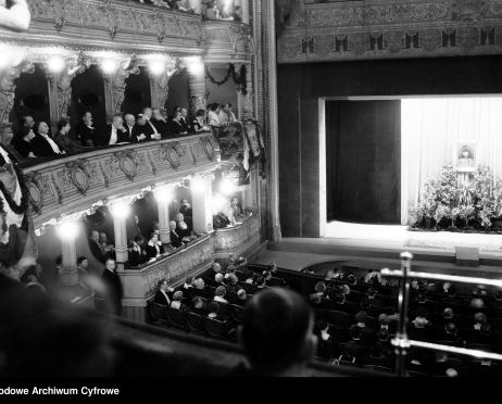 Na zdjęciu scena i widownia Dużej Sceny Teatru im. Juliusza Słowackiego w Krakowie. Na scenie znajduje się portret mężczyzny otoczony kwiatami. Jedna z lóż na pierwszym piętrze ozdobiona jest girlandami kwiatowymi. Widzowie ubrani są w odświętne, eleganckie ubrania. Zdjęcie czarno-białe.