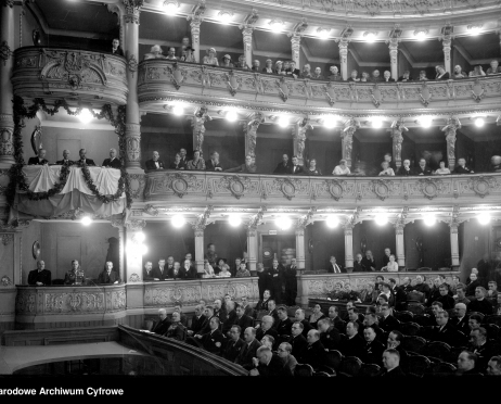Na zdjęciu widownia Dużej Sceny Teatru im. Juliusza Słowackiego w Krakowie. Jedna z lóż na pierwszym piętrze ozdobiona jest girlandami kwiatowymi. Widzowie ubrani są w odświętne, eleganckie ubrania. Zdjęcie czarno-białe.
