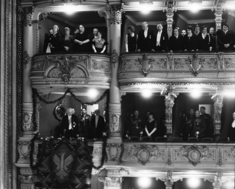 Na zdjęciu loże pierwszego i drugiego piętra widowni Dużej Sceny Teatru im. Juliusza Słowackiego w Krakowie. Jedna z lóż ozdobiona jest girlandami kwiatowymi. Widzowie ubrani są w odświętne, eleganckie ubrania. Wszyscy stoją jak do hymnu. Zdjęcie czarno-białe