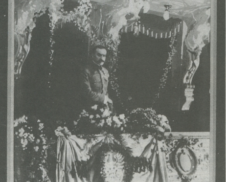 Na zdjęciu mężczyzna stoi w loży teatralnej. Loża ozdobiona jest kwiatowymi girlandami. Mężczyzna ma na sobie mundur wojskowy.