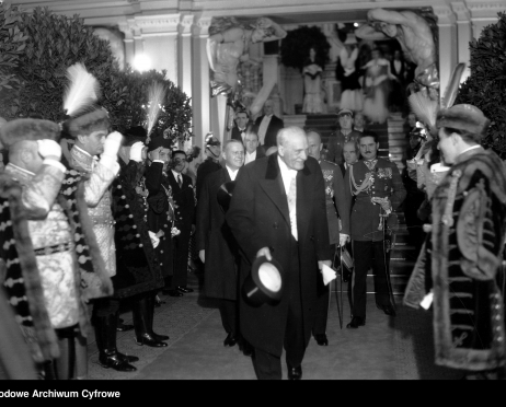 Na zdjęciu mężczyzna w podeszłym wieku przechodzi przez korytarz Teatru im. Juliusza Słowackiego w Krakowie. Po lewej i prawej stronie w rzędach stoją mężczyźni ubrani w historyczne stroje przypominające kontusz. Wszyscy salutują. Zdjęcie jest czarno-białe.