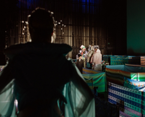 Na zdjęciu scena. Fragment scenografii do spektaklu. Na pierwszym planie głowa i tułów kobiety tyłem. Zamazany. ma rozłożone ręce. W oddali grupa ludzi stojących obok siebie. Rozmawiają między sobą. Na scenie rozłożone są parawany tak jak na plaży. Tło jest ciemne - zielone, szare, brązowe.