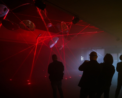 Na zdjęciu grupa osób przygląda się wiszącym u sufitu balonom w kształcie koła. Jest ciemno, ale w pomieszczeniu świecą czerwone światła dyskotekowe. Zgromadzone osoby robią zdjęcia instalacji.