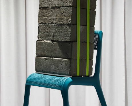Na zdjęciu niebieskie krzesło na którym ustawiono kilka szarych cegieł. Cegły związane są zielonymi tasiemkami. Krzesło stoi na białym podeście. Otoczone jest białą kurtyną.