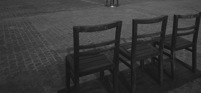 Zdjęcie przedstawia Plac Bohaterów Getta na Podgórzu w Krakowie. N pierwszym planie trzy krzesła. Ułożone koło siebie. W oddali plac na którym co kilka metrów stoją pojedyncze krzesła. Tłem są bloki, budynki. Zdjęcie jest czarno białe.