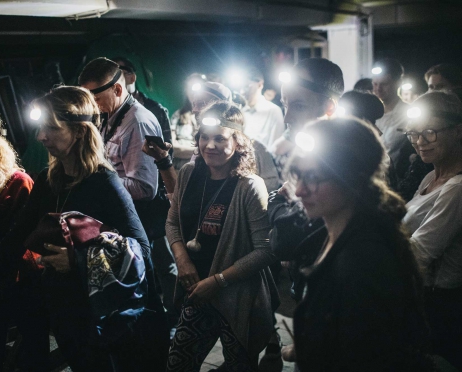 Na zdjęciu grupa ludzi stojąca w wąskim pomieszczeniu z niskim sufitem. Niektórzy mają na sobie latarki czołówki założone na głowie. Niektórzy trzymają przy sobie główke czosnku zawieszoną na sznurku. Pomieszczenie jest ciemne.