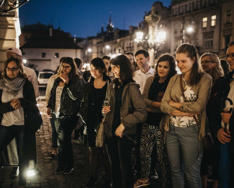 Na zdjęciu grupa ludzi stoi przed głównym wejściem do budynku teatru. Wszyscy wydają się spoglądać w jeden punkt. Jest pogodna noc, za ich plecami świecą latarnie miejskie. W tle budynki.