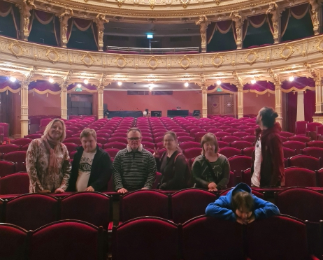 Zdjęcie zrobione na widowni Teatru im. J. Słowackiego. Widownia jest pusta, w jednym rzędzie krzeseł stoi 7 osób przodem do obiektywu. Wśród nich są osoby niskorosłe. Zdjęcie jakościowo jest lekko rozmazane.