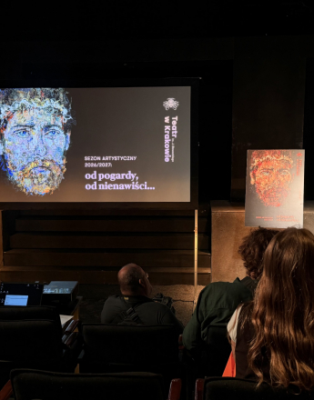 W przestrzeni Sceny Dom Machin na scenie stoi dyrektor Krzysztof Głuchowski, dyrektor Teatru imienia. Juliusza Słowackiego w Krakowie. Mężczyzna ma długie, siwe włosy i ubrany jest w jasną kurtkę dżinsową, jasne spodnie oraz czarną koszulkę z grafiką w barwach Ukrainy. niebieskiej i żółtej. Stoi przy mikrofonie na statywie, trzymając ręce przed sobą. Za nim znajduje się duży ekran projekcyjny, na którym wyświetlono grafikę przedstawiającą twarz Stanisława Wyspiańskiego, utrzymaną w odcieniach niebieskiego, pomarańczowego i żółtego. Po prawej stronie grafiki widnieje napis „Teatr w Krakowie” oraz „Sezon artystyczny 2026/2027: od pogardy, od nienawiści…”.Po obu stronach ekranu stoją plansze z grafikami promującymi sezon artystyczny, na jednej z nich widać obsadę aktorską. Pod ekranem znajdują się czarne schody prowadzące na scenę. W tle widoczne są ciemne ściany i czarna podłoga. Na pierwszym planie, tyłem do obiektywu, siedzą osoby z publiczności na teatralnych fotelach.