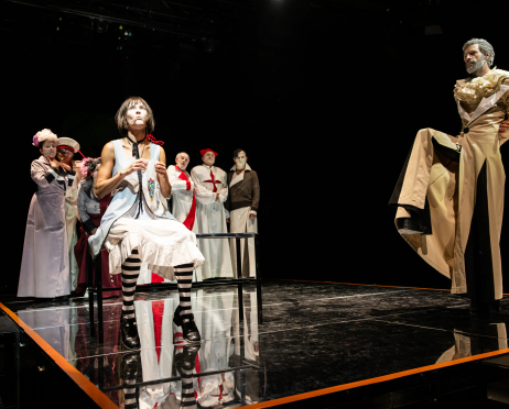 Scena ze spektaklu „O!peretka” Teatru imienia Stanisława Ignacego Witkiewicza w Zakopanem. Na czarnej, lśniącej scenie znajduje się grupa aktorów w charakterystycznych, groteskowych kostiumach. Na pierwszym planie, po lewej stronie, siedzi kobieta w białej sukni bez rękawów, z czarną wstążką pod szyją i czarno-białymi, pasiastymi pończochami. Siedzi na metalowej konstrukcji i trzyma w dłoniach niewielki przedmiot. Po prawej stronie sceny stoi mężczyzna w jasnym, obszernym kostiumie z dużym, ozdobnym kołnierzem. Wykonuje dynamiczny gest, unosząc jedną nogę wysoko i opierając ją na metalowej konstrukcji. Za kobietą znajduje się grupa aktorów ubranych w białe kostiumy z czerwonymi elementami. Część postaci ma na głowach fantazyjne nakrycia głowy. Scena jest niemal całkowicie pozbawiona dekoracji, a czarne tło i lśniąca powierzchnia podłogi tworzą kontrast z jasnymi kostiumami aktorów.