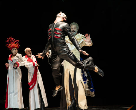 Scena ze spektaklu „O!peretka” Teatru imienia Stanisława Ignacego Witkiewicza w Zakopanem. Na ciemnej scenie występuje pięć postaci w wyrazistych, teatralnych kostiumach. Na pierwszym planie aktor ubrany w czarny, obcisły kostium z czerwonymi rozcięciami na piersi wykonuje dynamiczny ruch z uniesioną głową i szeroko rozstawionymi rękami, jedną nogę ma wysoko uniesioną w powietrzu. Za nim stoi aktor w jasnym, długim kostiumie z charakterystycznym jasnym kołnierzem. Unosi dłoń w geście zatrzymania i spogląda na znajdującą się przed nim postać. Po lewej stronie sceny stoją dwie postacie w długich, białych kostiumach z czerwonymi elementami. Jedna z nich ma na głowie fantazyjną, czerwoną konstrukcję przypominającą płomienie. Czarna przestrzeń sceniczna stanowi tło dla jasnych i intensywnie kolorowych kostiumów.