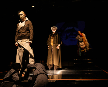 Scena ze spektaklu „O!peretka” Teatru imienia Stanisława Ignacego Witkiewicza w Zakopanem. Na niemal całkowicie zaciemnionej scenie, oświetlonej punktowymi reflektorami, znajdują się trzy postacie w wyrazistych kostiumach.Na pierwszym planie po lewej stronie stoi mężczyzna w ciemnej, dopasowanej marynarce z jasnym, szerokim kołnierzem oraz jasnych, szerokich spodniach. Ma poważny wyraz twarzy. W centrum sceny stoi postać w długim, ciemnym płaszczu, kapeluszu i jasnej kamizelce. W głębi, po prawej stronie, znajduje się postać w rozbudowanym, kolorowym kostiumie z dużą dekoracją z piór. Na pierwszym planie, u stóp podestu, znajdują się dwie osoby ubrane na czarno. Jedna klęczy i pochyla się nad podłogą, druga leży lub jest pochylona nisko przy krawędzi sceny. Aktorzy znajdują się na czarnej, wielopoziomowej konstrukcji scenicznej, której powierzchnia odbija światło. W tle widoczna jest duża, ciemna projekcja lub element scenografii w odcieniach granatu.