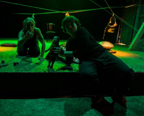 Kobieta siedzi na schodku, porusza drewnianą lalką. Obok niej klęczy druga kobieta. Wskazuje palcem przed siebie. Przestrzeń oświetlona jest zielonym światłem. Tło jest czarne.