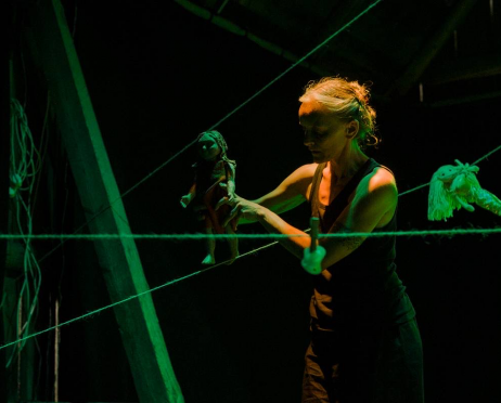 Kobieta stoi wśród pajęczyny ze sznurków. Po jednym z nich przesuwa drewnianą lalkę. Przestrzeń jest ciemna, świetlona jest zielonym światłem. Tło jest czarne.