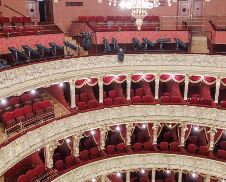 Zdjęcie zrobione z górnego piętra na loże na widowni Teatru Słowackiego. Ujęcie na 3 piętra lóż i fragment widowni. Krzesła w kolorze bordowym, filary bogato zdobione. Z sufitu zwisa ogromny rozświetlony, rzeźbiony żyrandol. Widownia jest pusta.