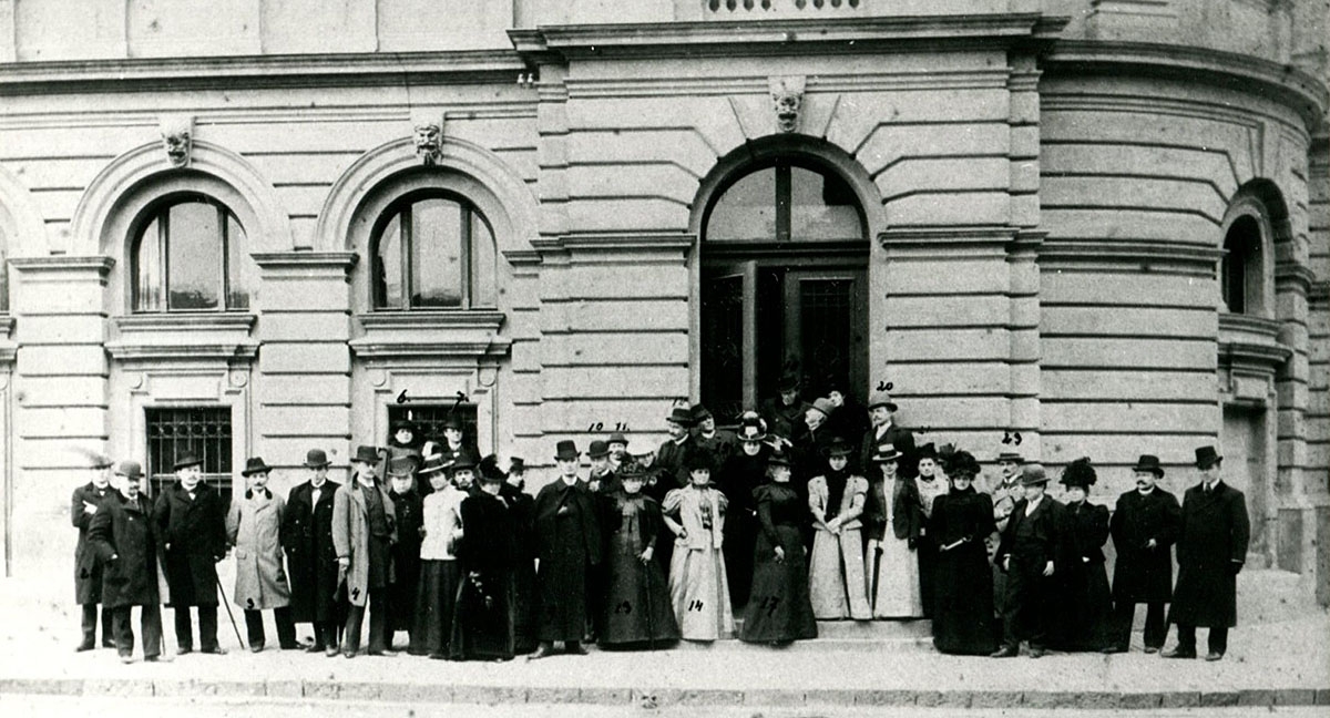 Zdjęcie zrobione z oddali. W centralnej części zdjęcia fragment budynku głównego Teatru Słowackiego. Przed budynkiem grupa ludzi stoją ustawieni jakby do zdjęcia. Jest to zespół aktorski z roku 1893 roku. Patrzą prosto przed siebie. Kobiety mają długie suknie, kapelusze. Mężczyźni garnitury oraz cylindry lub kapelusze. Zdjęcie jest czarno białe.