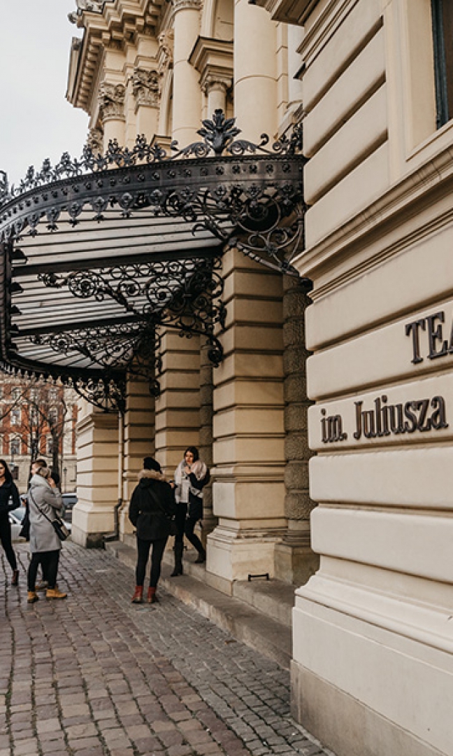 Na zdjęciu wejście do budynku Teatru im. Juliusza Słowackiego w Krakowie. Budynek jest koloru żółtego. Nad drzwiami wejściowymi jest niewielki, czarny, zdobiony daszek. Przed wejściem stoi niewielka grupa ludzi ubranych w ciepłe kurtki.
