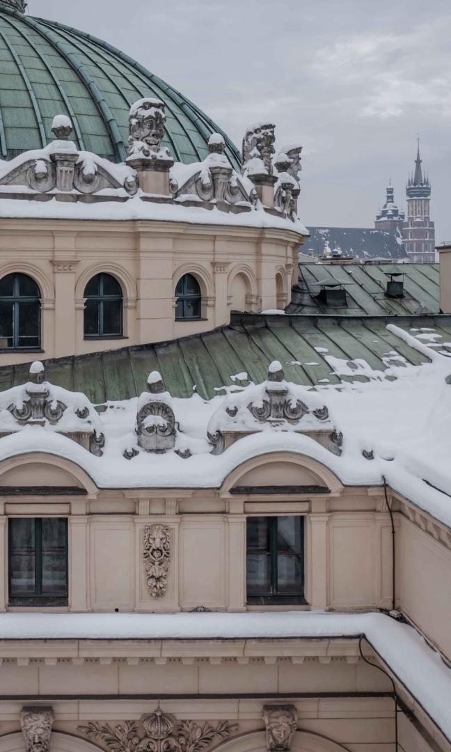 Zdjęcie fragmentu budynku Teatru Słowackiego w Krakowie zrobione na wysokości dachu. Dach jest pokryty śniegiem. w tle widoczne wieże kościoła Mariackiego. Niebo jest szare.