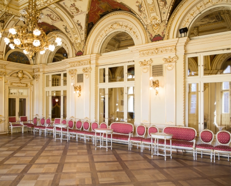 Zdjęcie foyer Teatru Słowackiego. Bogato zdobiona sala, z drewnianym parkietem, ściany zdobione złotem, na suficie malowane freski. Wzdłuż ściany z oknami stoją stylowe krzesła z bordowymi obiciami. Z sufitu zwisa mosiężny żyrandol, z kloszami w kształcie kwiatów.