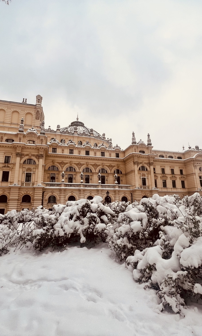 Zdjęcie secesyjnego gmachu Teatru im. Juliusza Słowackiego w Krakowie. Teatr jest w kolorze kremowym. Ujęcie zimą, na boczną ścianę budynku. U dołu zdjęcia na krzakach leży śnieg, niebo jest szare.