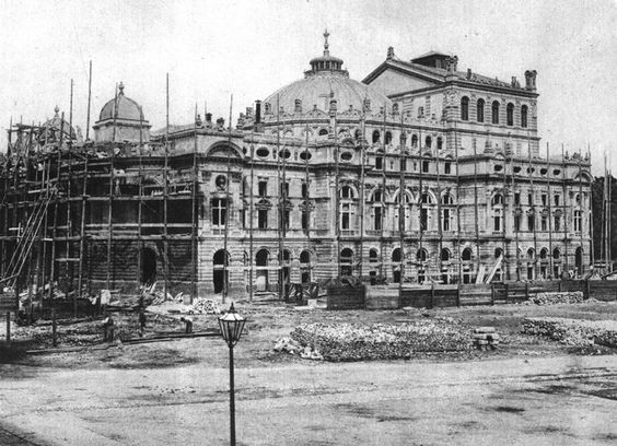 Na zdjęciu budynek Teatru Słowackiego zrobiony w oddali. Budynek jest w trakcie budowy. Są rusztowania przy ścianach. Odkopane fundamenty. Zdjęcie jest czarno białe. Powierzchnia wokół budynku jest pusta. W tle chmury.