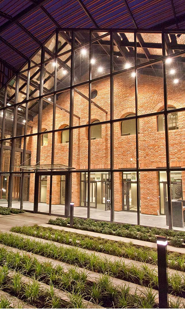 Zdjęcie budynku Małopolskiego Ogrodu Sztuki. Budynek jest z czerwonej cegły. Przed ścianą dobudowane pomieszczenie przypominające oranżerię z szyb i czarnej stali. Ściana budynku jest oświetlona. Przed wejściem rabaty z zielonymi roślinami. Zdjęcie wykonane nocą