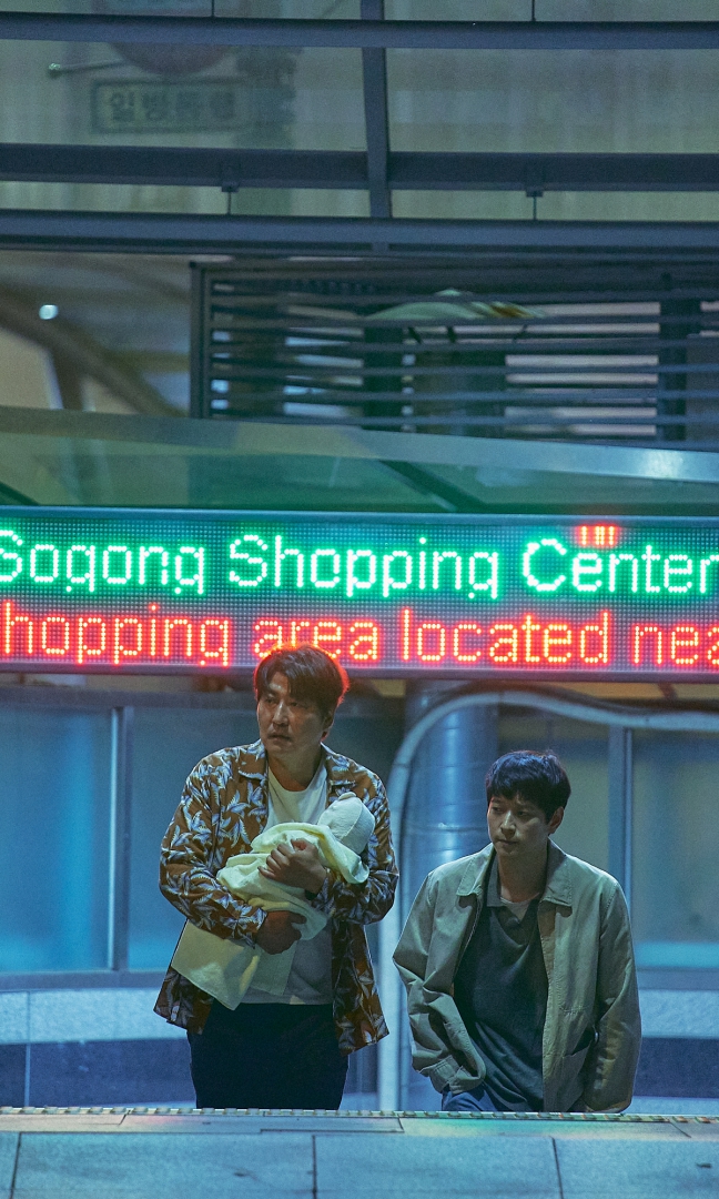 U dołu zdjęcia dwie głowy azjatyckich mężczyzn, patrzą w bok, wyglądają na zmęczonych. Nad nimi neon z napisami zielonymi i czerwonymi. W tle metalowa konstrukcja budynku.