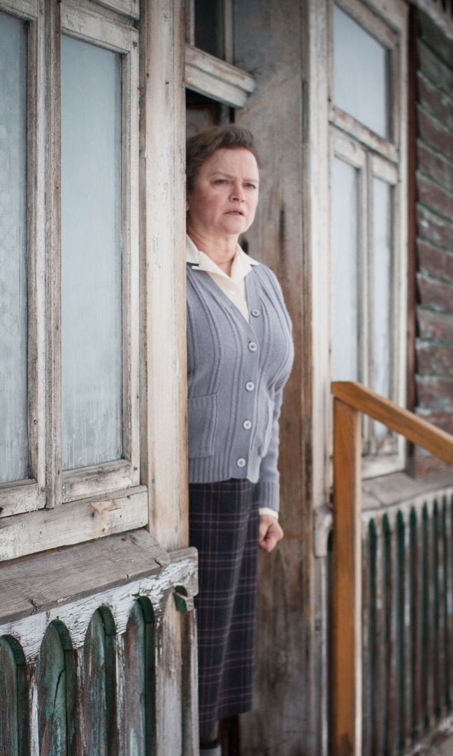 Zdjęcie kobiety w średnim wieku. Stoi na progu domu który ma obdrapane ściany i okna. Ubrana w fioletowy zapinany sweterek spod którego wystaje biały kołnierzyk. Ma smutną, lekko przerażoną twarz, wygląda na zmęczoną.