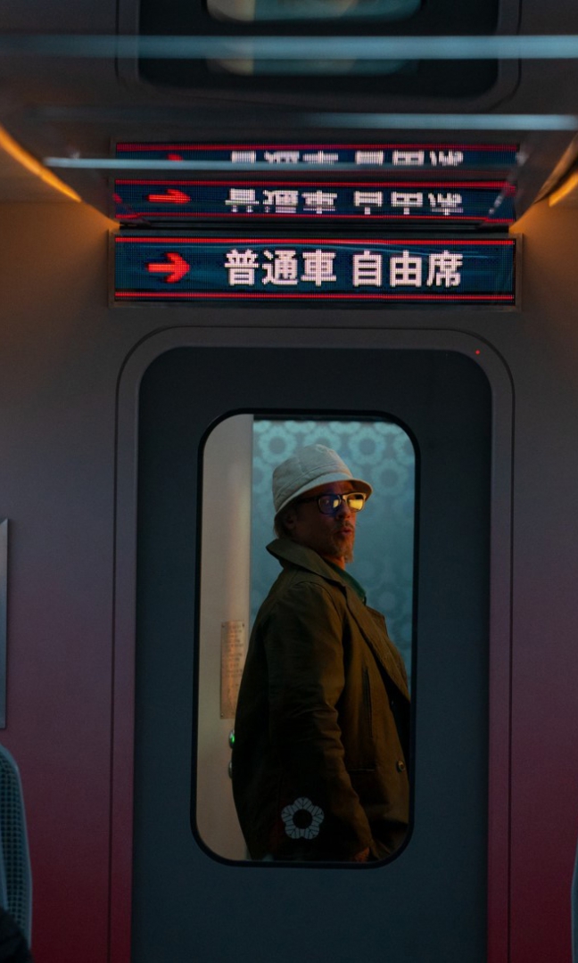 Na zdjęciu mężczyzna ubrany w kapelusz, płaszcz i okulary. Stoi w drzwiach do następnego przedziału pociągu. Nad jego głową wiszą tablice z napisami w języku japońskim.