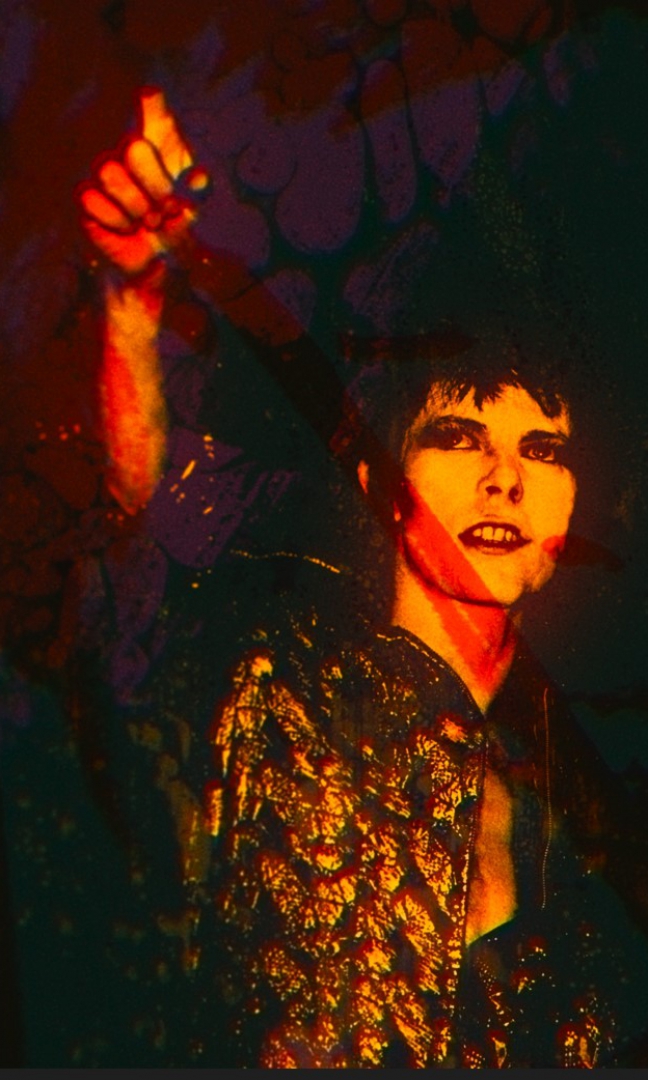 Przekoloryzowane zdjęcie młodego mężczyzny, stylizowanego na lata 80-te. Ma mocny makijaż, krótkie ciemne włosy z pazurkami, błyszczący strój. Widać zęby, podnosi rękę w górę wskazując palcem.