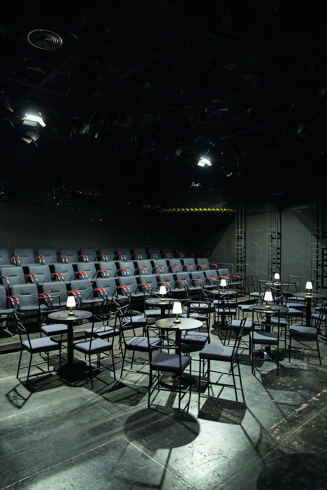 Zdjęcie sceny i widowni Domu Machin w Krakowie. W czarnej przestrzeni teatralnej na scenie rozłożone są stoliki i krzesła. Za nimi kilka rzędów foteli na widowni.