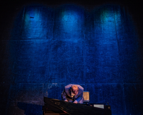 Zdjęcie robione z góry. Na zdjęciu w centralnej części fragment czarnego fortepianu. Przy fortepianie siedzi młody mężczyzna. Ubrany jest w granatowy garnitur. Gra. Tłem jest drewniana sceniczna podłoga, która oświetlona jest na kolor niebieski.