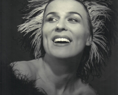 Zdjęcie to czarno-biały portret aktorki Marty Stebnickiej. Młoda kobieta ma na głowie perukę przypominającą postrzępione puchowe piórka. Kobieta uśmiecha się radośnie i spogląda w lewo poza kadr. Na jej lewym ramieniu opada czarny szal boa.