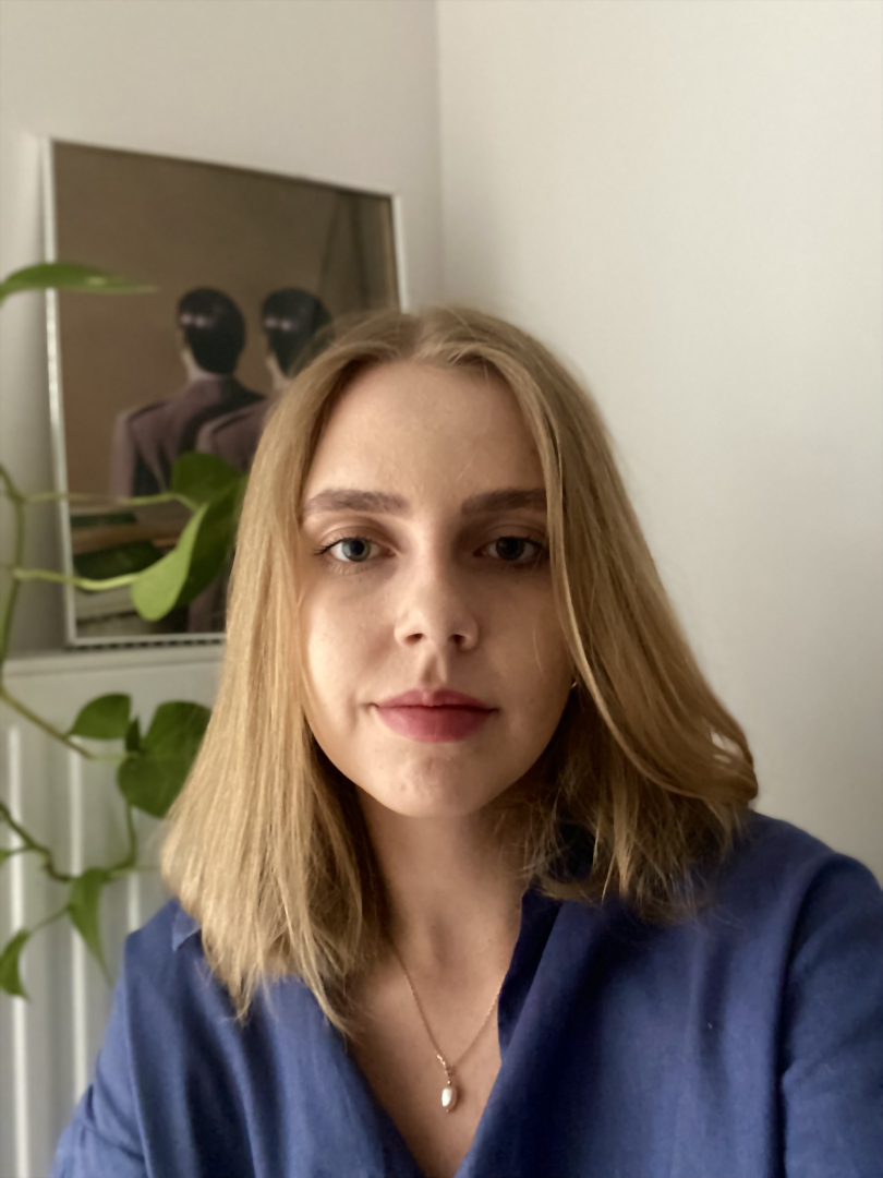 Portretowe zdjęcie Julii Butryn. 26-letnia kobieta o jasnej cerze i blond włosach patrzy wprost w obiektyw. Ma na sobie granatową koszulę oraz naszyjnik z perłą. W tle widoczna jest jasna ściana, zielona roślina doniczkowa oraz obraz przedstawiający dwie postacie na ciemnym tle. Fotografię wykonano w miękkim, naturalnym świetle.