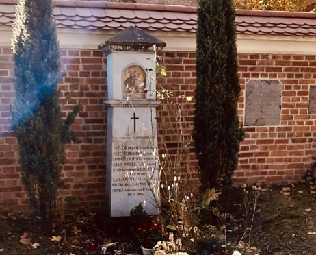 Na zdjęciu kapliczka z grobu Lucjana Rydla, Kapliczka jest wąska, podłużna, pomalowana na biało. Stoi na tle muru z czerwonej cegły. Po jej dwóch stronach rosną tuje. Przed kapliczką róża, suche liście na ziemi. Pomiędzy nimi stoją znicze.
