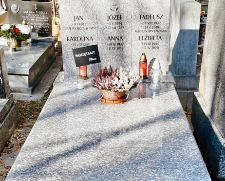 Zdjęcie grobu rodziny Szybowskich na cmentarzu. Nagrobek jest kamienny. W tle inne groby. Na płycie nagrobka stoją znicze, wieniec i czarna chorągiewka z napisem PAMIĘTAMY i z logotypem Teatru Słowackiego w Krakowie.
